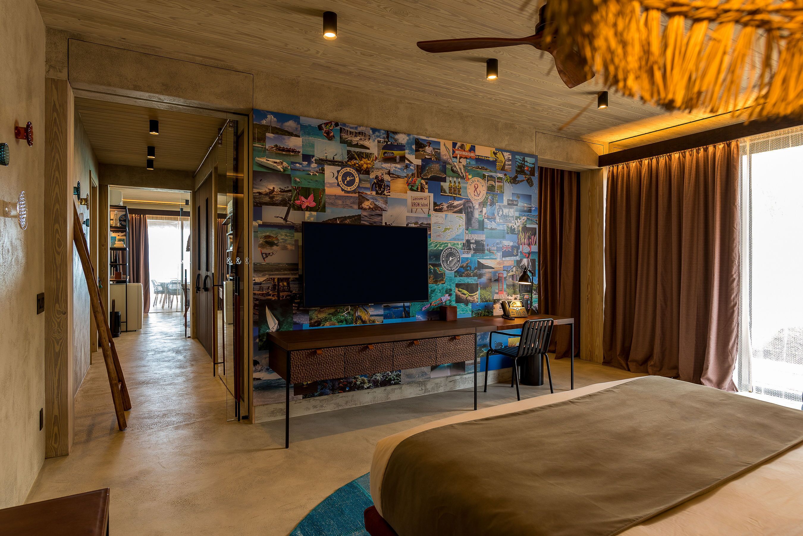Een moderne hotelkamer met een groot bed, een flatscreen televisie aan de wand met een fotocollage, een bureau met stoel en een groot raam met gordijnen.