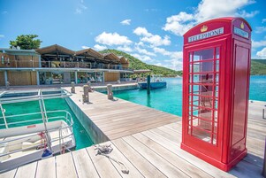 Cabina telefónica roja en un muelle de madera, con una piscina y barcos.