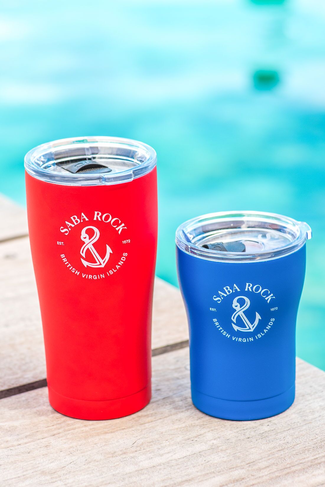 Twee Saba Rock drinkbekers, rood en blauw, op een houten ondergrond in de buurt van een zwembad.