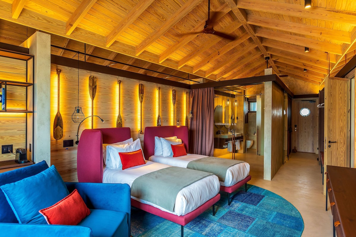 Habitación de hotel moderna con vigas de techo de madera, dos camas con ropa de cama roja y blanca, un sofá azul y una alfombra azul estampada.
