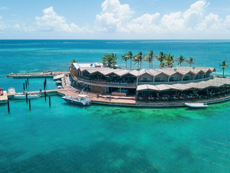 A tropical island resort with multiple overwater bungalows connected by walkways, a dock with boats, and clear turquoise waters.