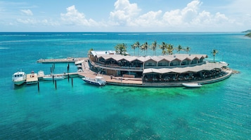 Un resort isleño tropical con varias cabañas sobre el agua conectadas por pasarelas, un muelle con barcos y aguas turquesas cristalinas.