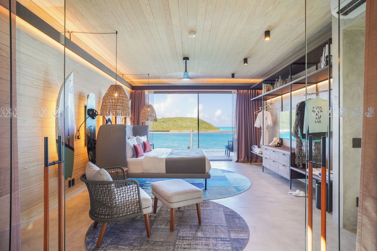 Un dormitorio moderno con una cama grande, una zona de estar con una silla y reposapiés, un armario y vistas al océano.