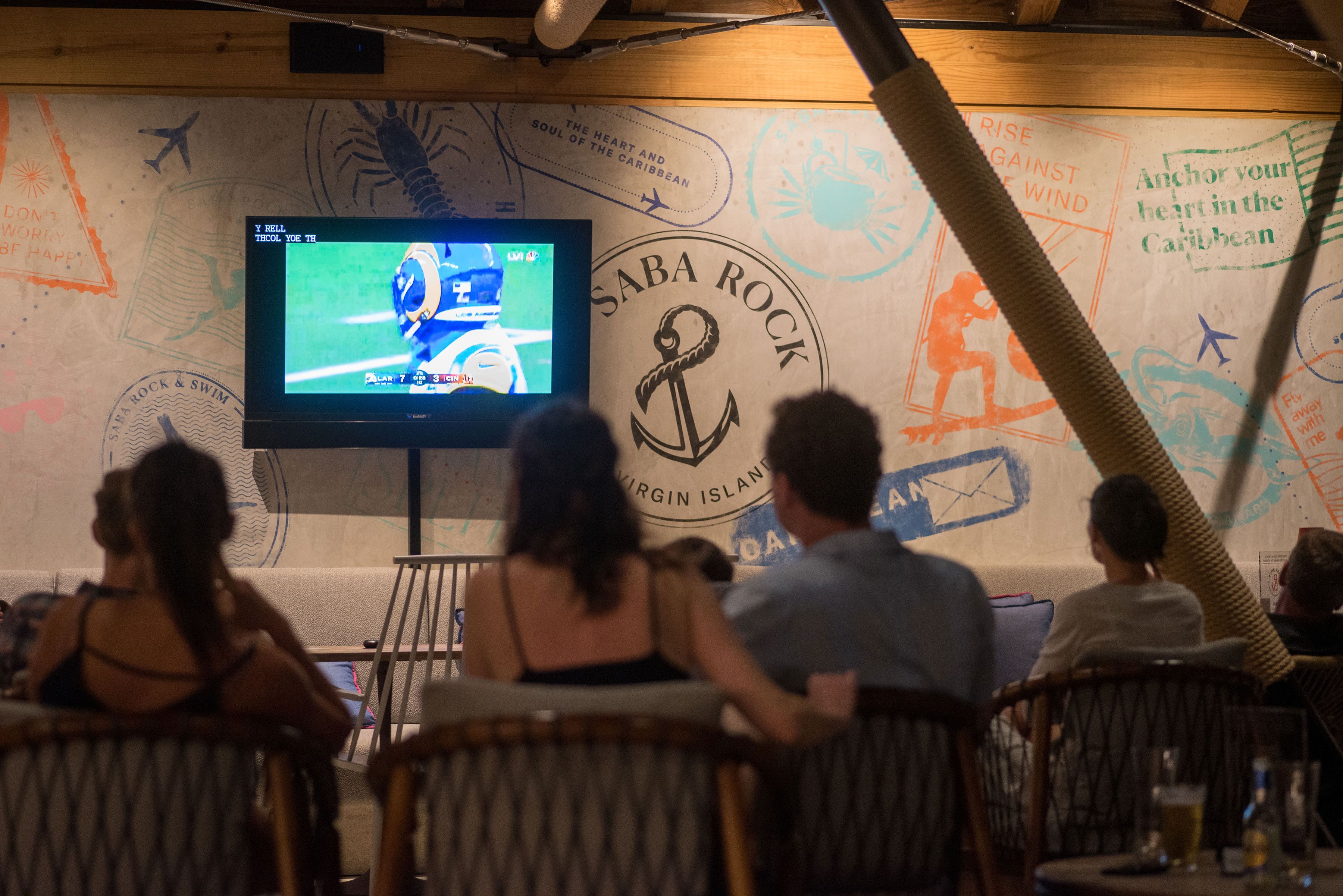 Un restaurante con una gran pantalla mostrando un evento deportivo, paredes de madera con decoración náutica y clientes sentados en mesas.