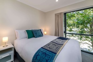 A hotel room with a bed, bedside lamps, and a window with a view of trees.