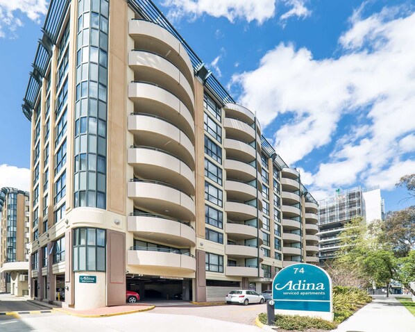 A multi-story serviced apartment building with a curved facade, a parking garage entrance, and a clear sky.