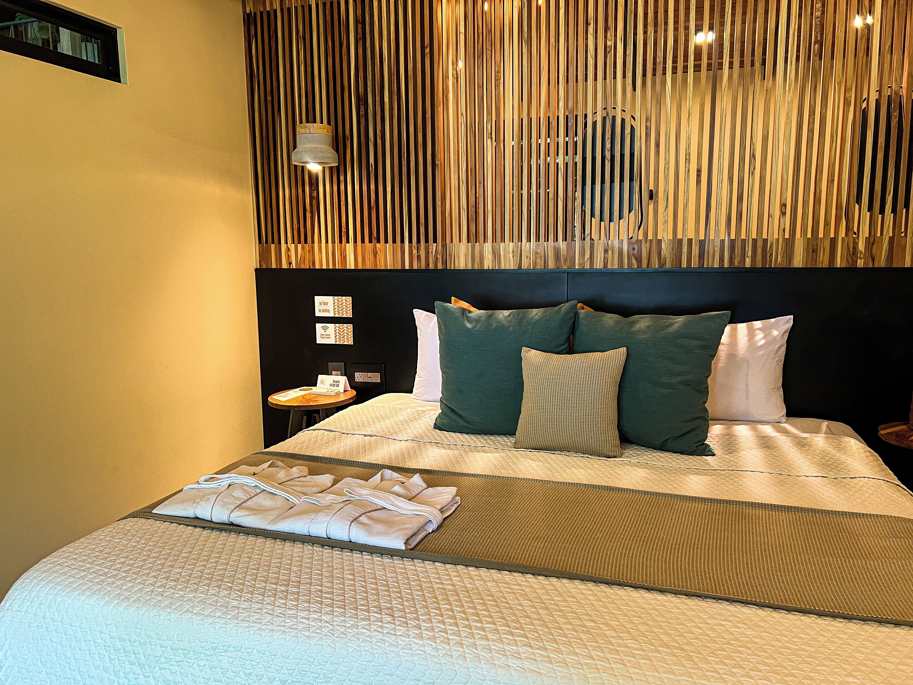 Una cama bien hecha con almohadas, una mesita de noche con un libro y un teléfono, y una pared con listones de madera verticales y una lámpara colgante.