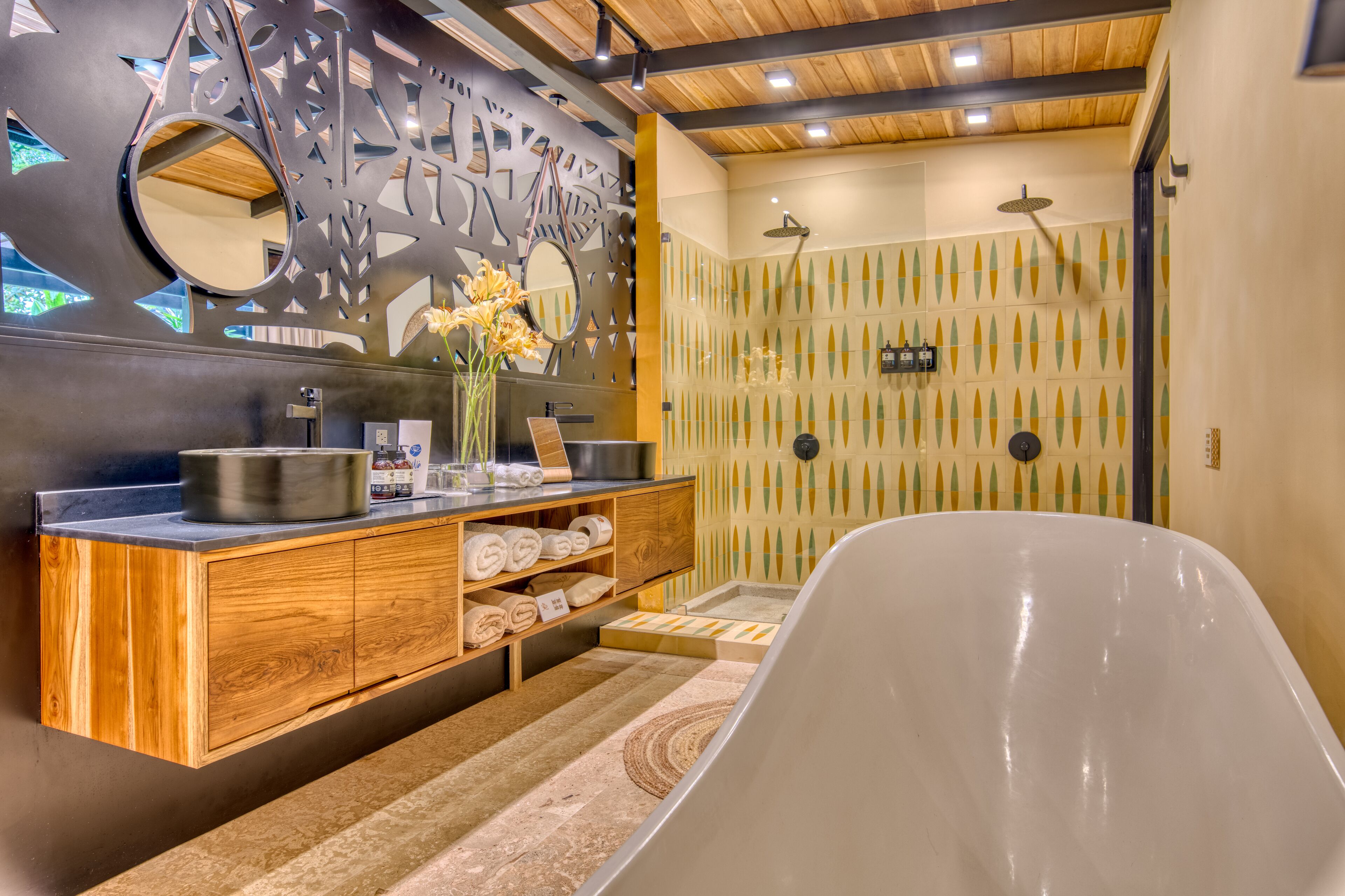 A modern bathroom with a wooden vanity, a large mirror, and a shower area with a unique geometric tile design.