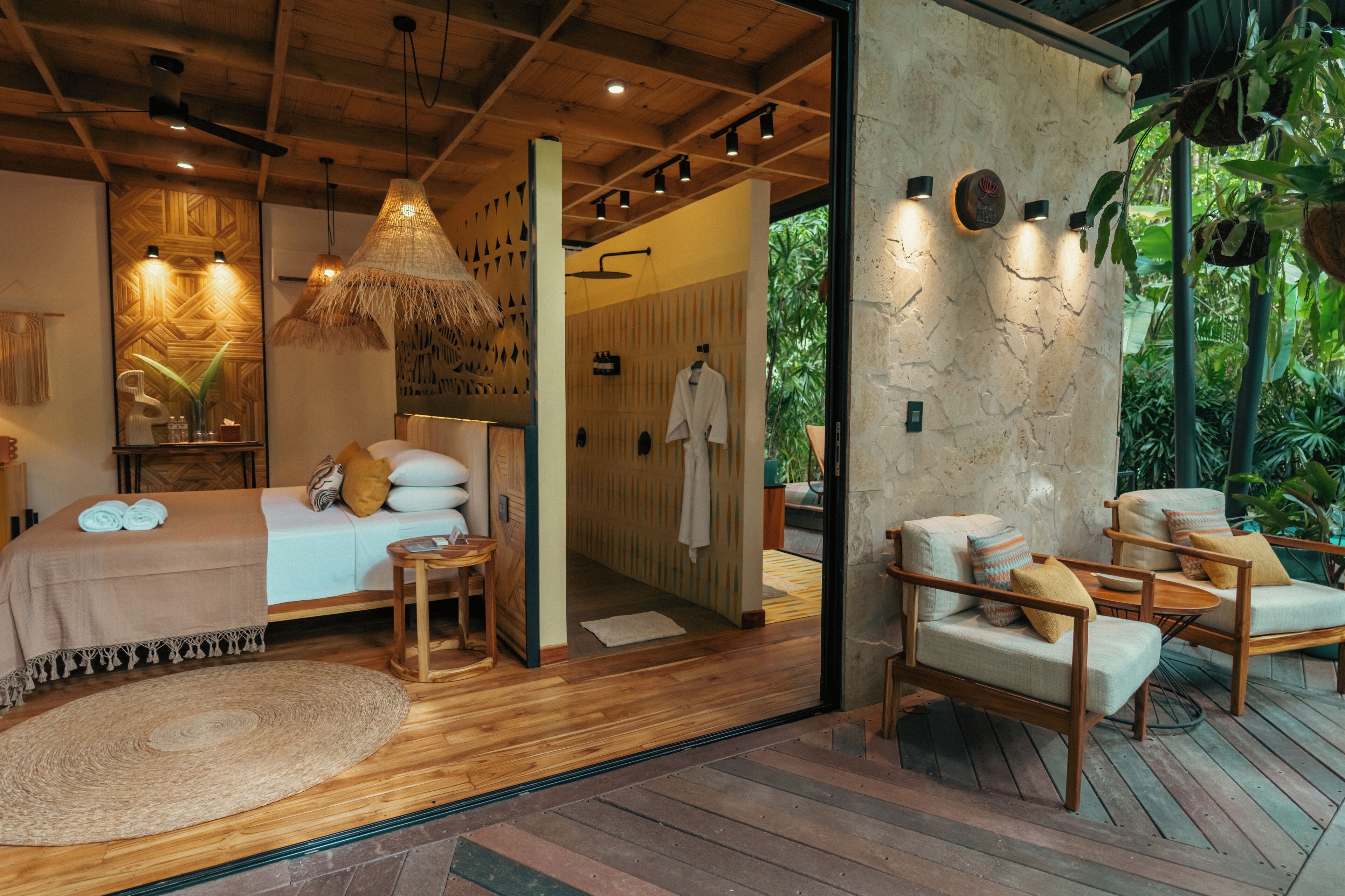 Une chambre moderne avec un plafond en bois, un grand lit recouvert d’une courtepointe beige, une suspension lumineuse en fibres tressées et une vue sur un jardin luxuriant.