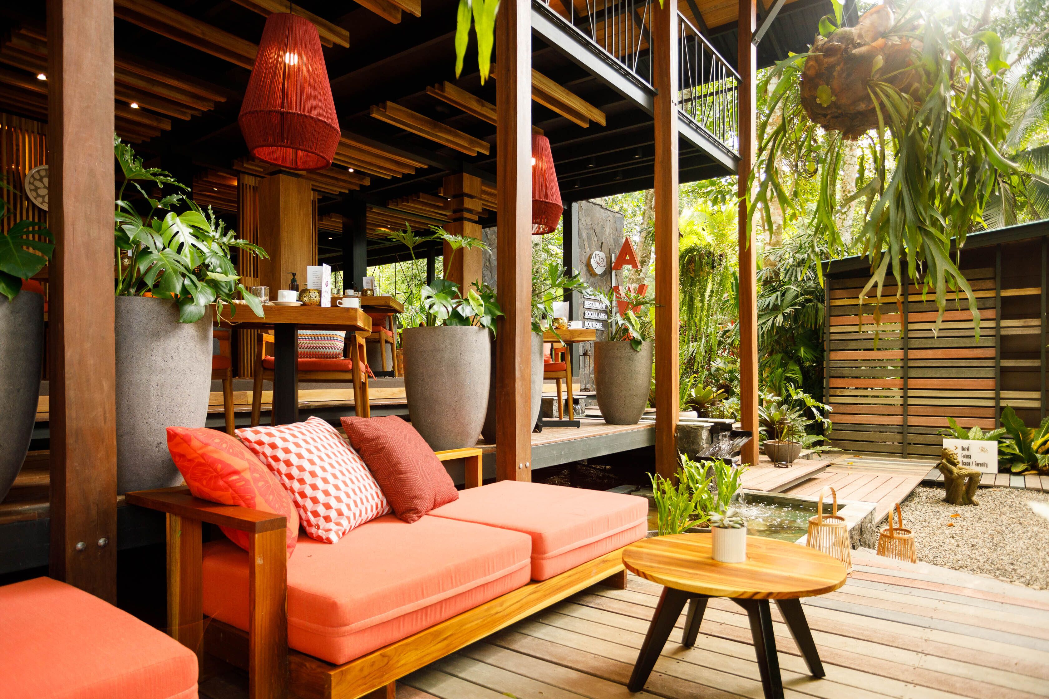 A wooden deck with a sofa and a table, surrounded by large potted plants and a small water feature.