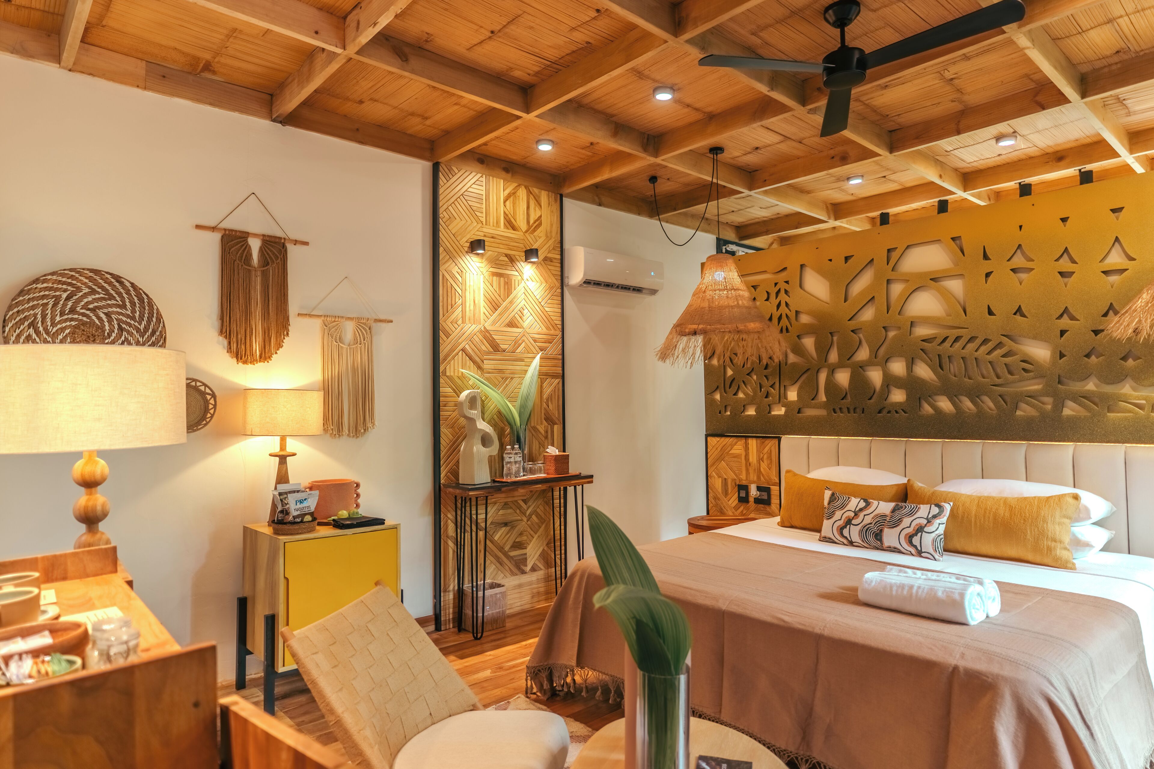 Quarto bem iluminado com teto de madeira, cama com travesseiros brancos e amarelos, mesa de cabeceira com abajur e painel decorativo na parede.
