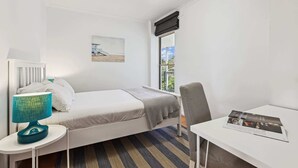 Ein kleines, aufgeräumtes Schlafzimmer mit einem Einzelbett, einem Schreibtisch, einem Stuhl, einer Lampe und einem Fenster mit Blick auf Grünflächen.