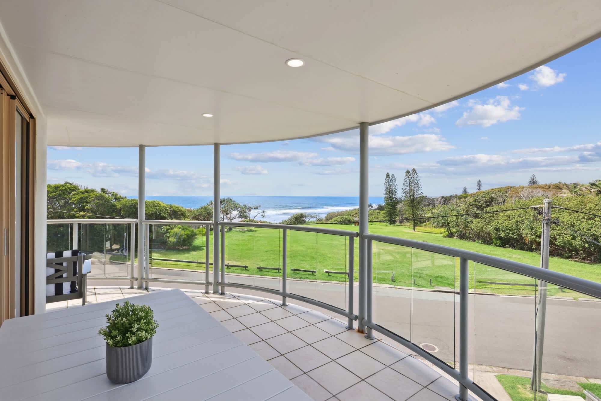 Un balcon doté d’une balustrade en verre incurvée, d’une plante en pot et offrant une vue sur un paysage verdoyant et l’océan.
