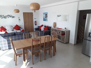 A modern living room with a sofa set, a dining table with chairs, a kitchen area with a refrigerator, and a staircase leading to another level.