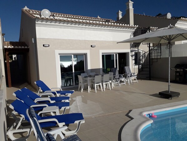 A poolside area with lounge chairs, a dining table, and a barbecue grill.