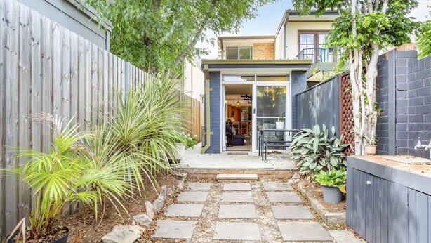 A narrow stone pathway leading to a modern house with a wooden fence on one side and a brick wall on the other.