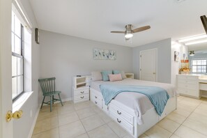 Quarto com cama, cadeira, mesa de cabeceira, cômoda, ventilador de teto e janela.
