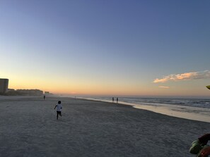 Une plage où l’on voit une personne qui se promène, des bâtiments en arrière-plan et un ciel dégagé.