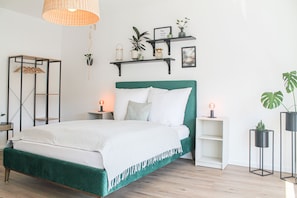 A neatly arranged bedroom with a green upholstered bed, white bedding, and two bedside tables with lamps. There are wall-mounted shelves with plants and decorative items.