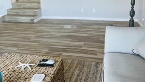 Ein Wohnzimmer mit Holzboden, einem grauen Sofa, einem gewebten Couchtisch und einem Fernseher, der eine Strandansicht zeigt.