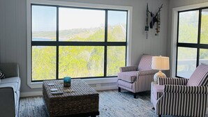 Ein geräumiges Wohnzimmer mit großen Fenstern, einem gewebten Couchtisch, gestreiften Sessel und einem grauen Sofa.