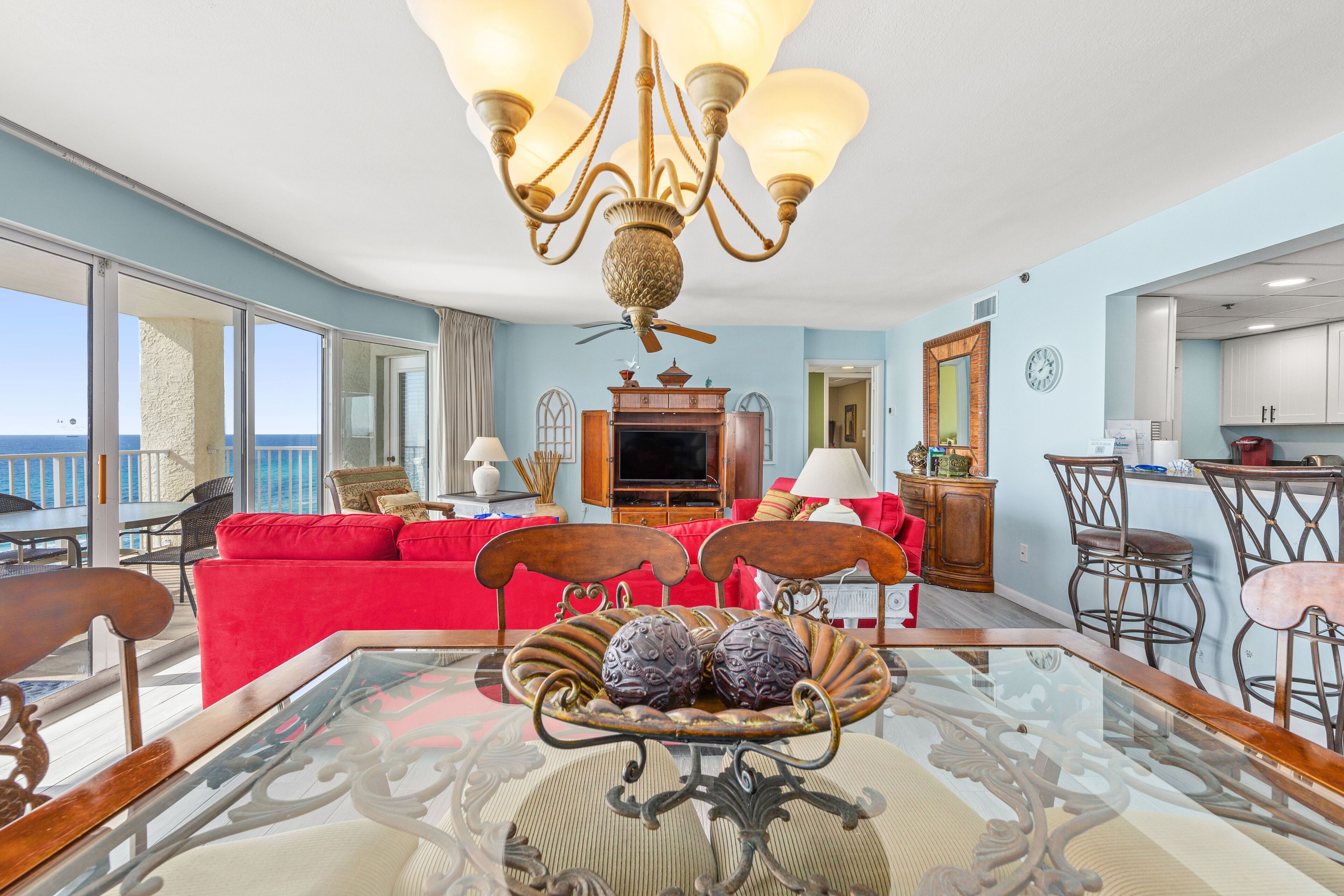 A spacious living room with a red sofa, a glass coffee table with decorative items, a chandelier, and a view of the ocean.