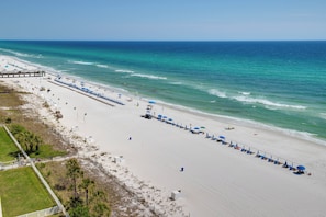 A beach with a long row of umbrellas and lounge chairs, a pier, and a clear view of the ocean.