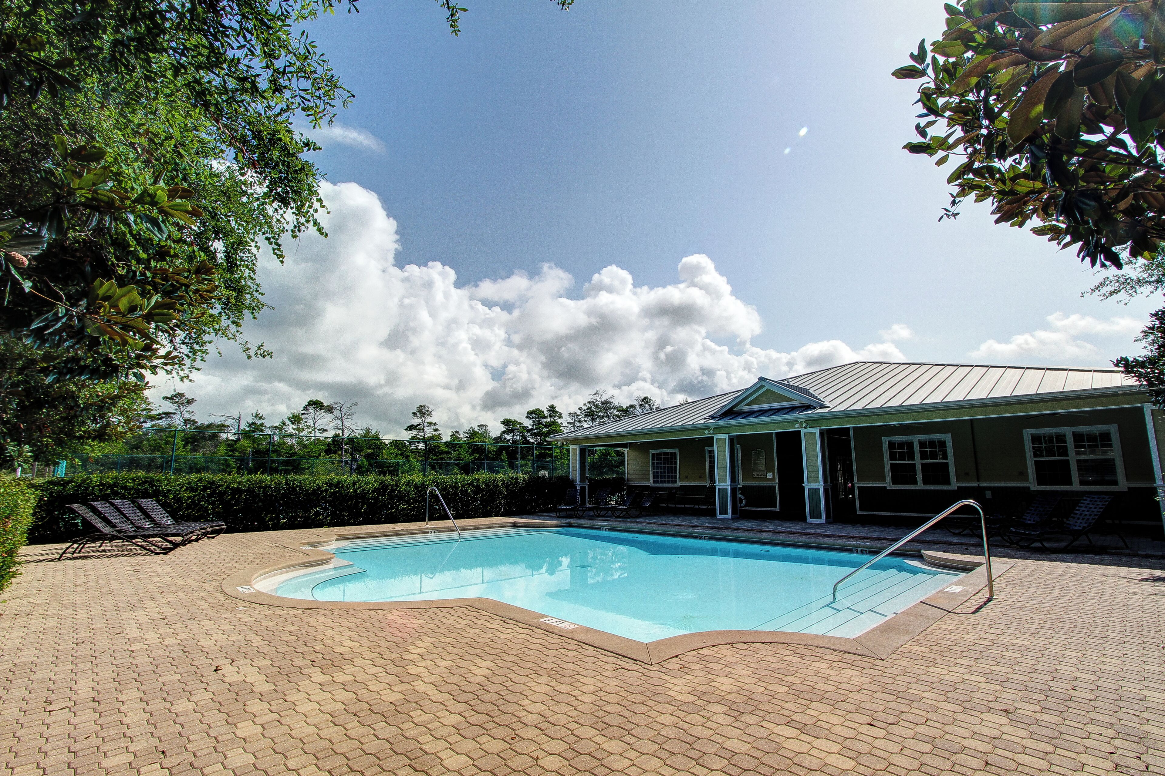 A swimming pool with a brick deck, lounge chairs, and a building with a green roof in the background.