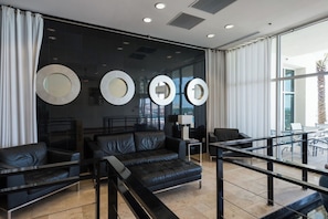 A modern hotel lobby with a black leather sofa, a glass coffee table, and a dining area with a view of palm trees. - Sterling Breeze 1105 Sandy Haven - Sleeps 6! Gulf Views! Near Pier Park! (Panama City Beach)