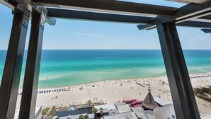 A beach view from a high vantage point, featuring a sandy beach, clear turquoise waters, and a few buildings with a thatched-roof structure.