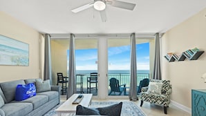 Una sala de estar con un sofá gris, un sillón estampado, una mesa de centro y un aparador azul turquesa. Hay un balcón con vistas al océano.