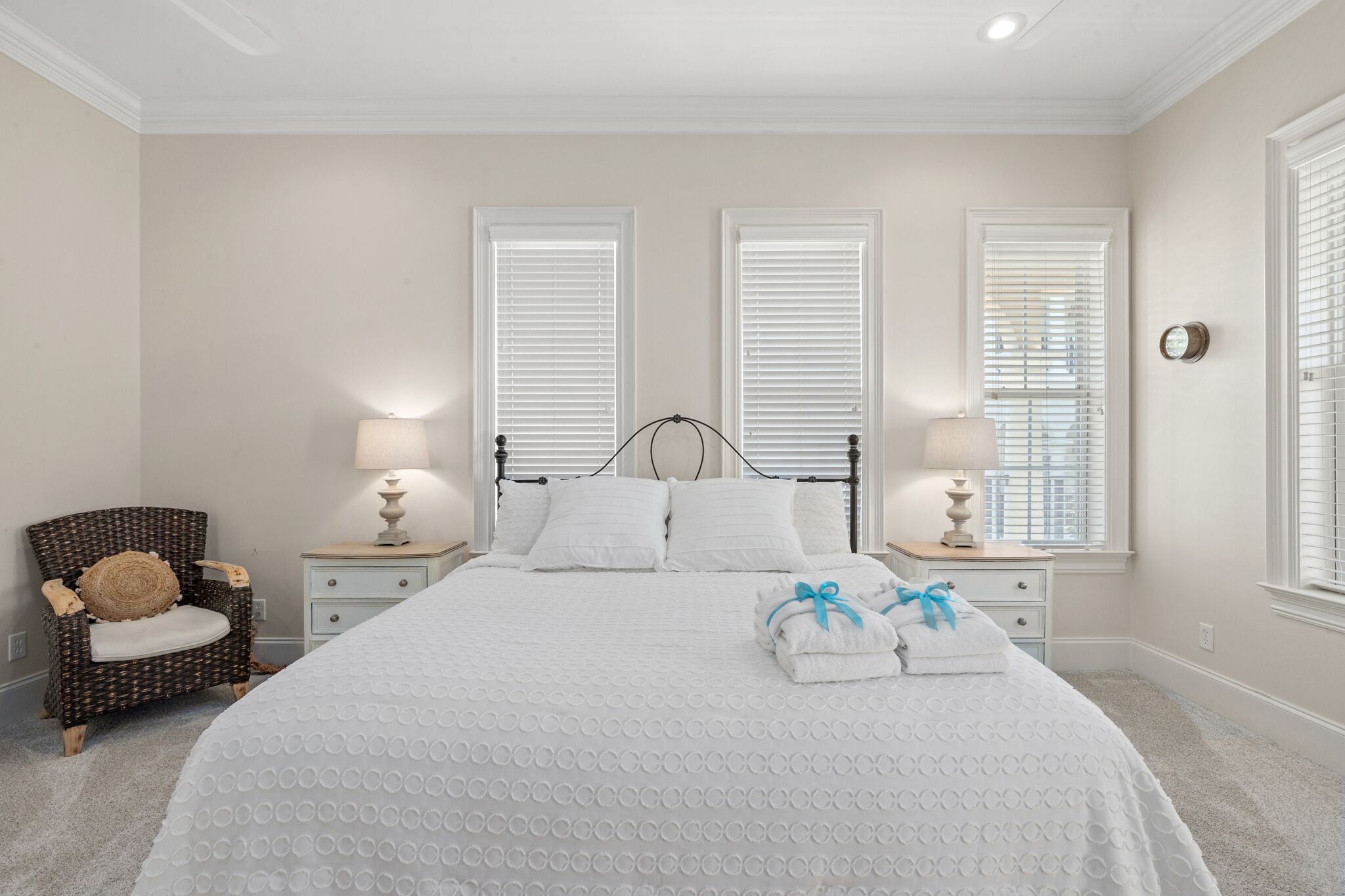 A neatly arranged bedroom with a large bed, two bedside tables with lamps, a wicker chair, and three windows with blinds.