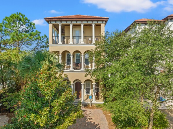 A two-story building with a balcony, columns, and a paved entrance surrounded by lush greenery. - Luxury Home Steps From Beach in Gated Community (Santa Rosa Beach)