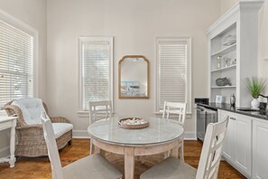 A dining area with a round glass table, white chairs, a wicker chair, and a white cabinet with shelves. - Luxury Home Steps From Beach in Gated Community (Santa Rosa Beach)