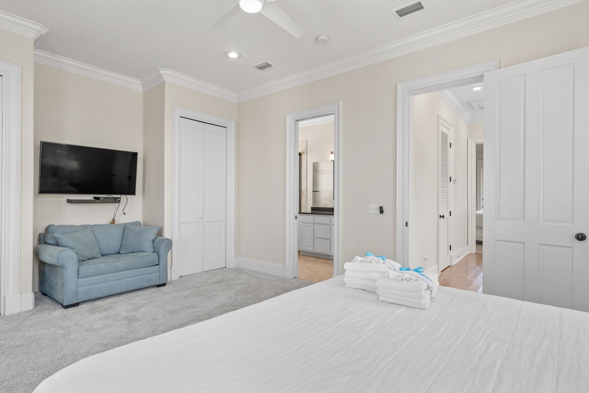 A bedroom with a large bed, a blue sofa, a TV, and a kitchen visible through an open door.