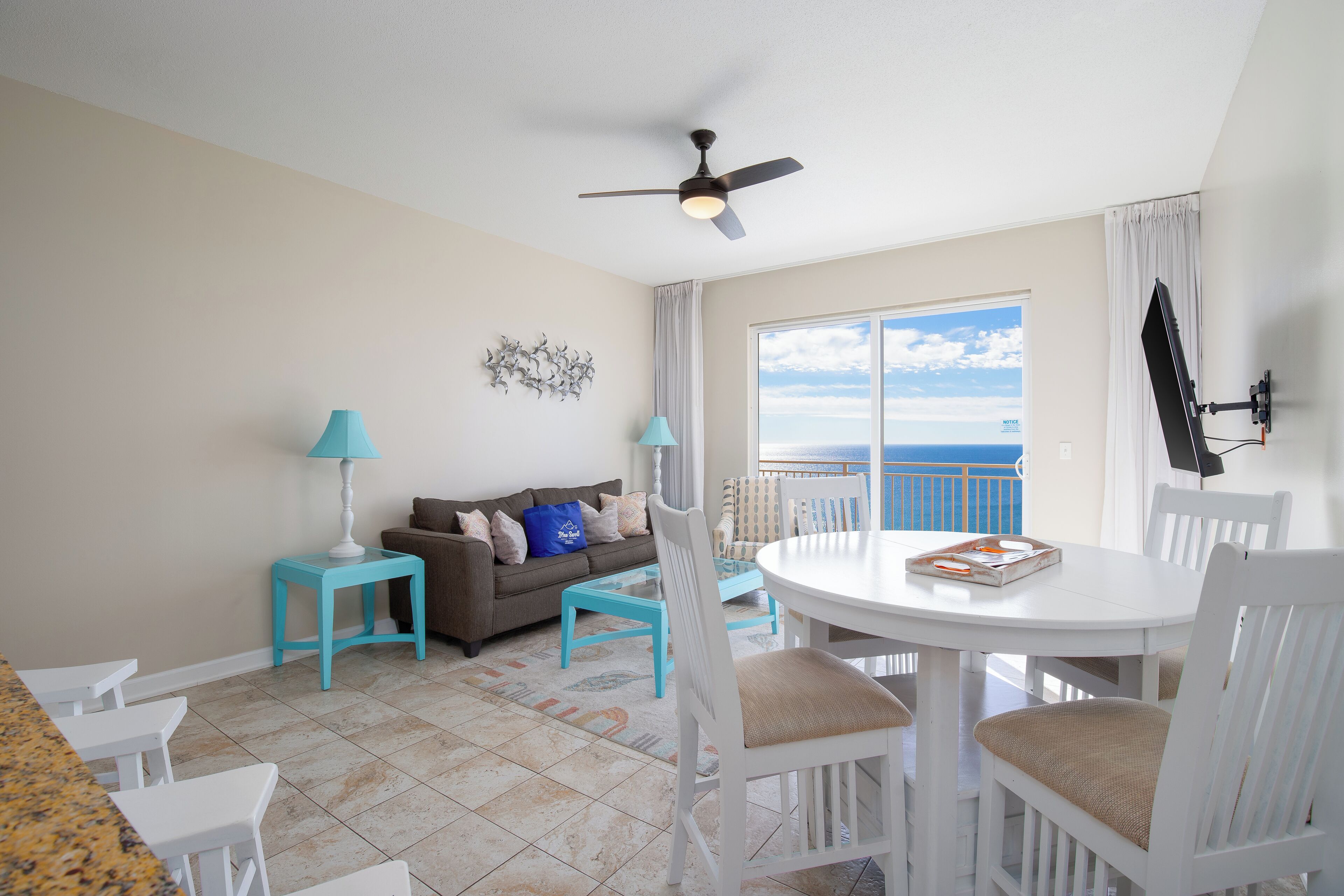 A room with a balcony, a ceiling fan, a dining table with chairs, a sofa, and a TV mounted on the wall.