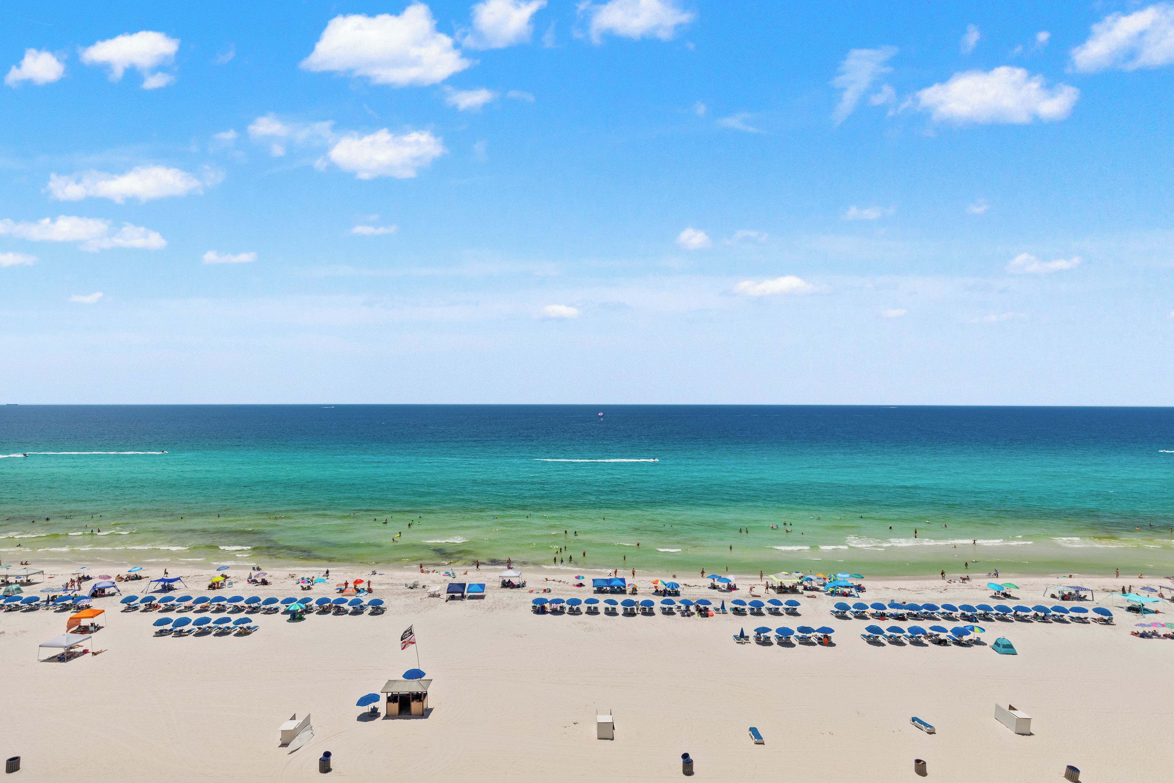 A beach with rows of sun loungers and umbrellas, a lifeguard tower, and a clear view of the ocean.