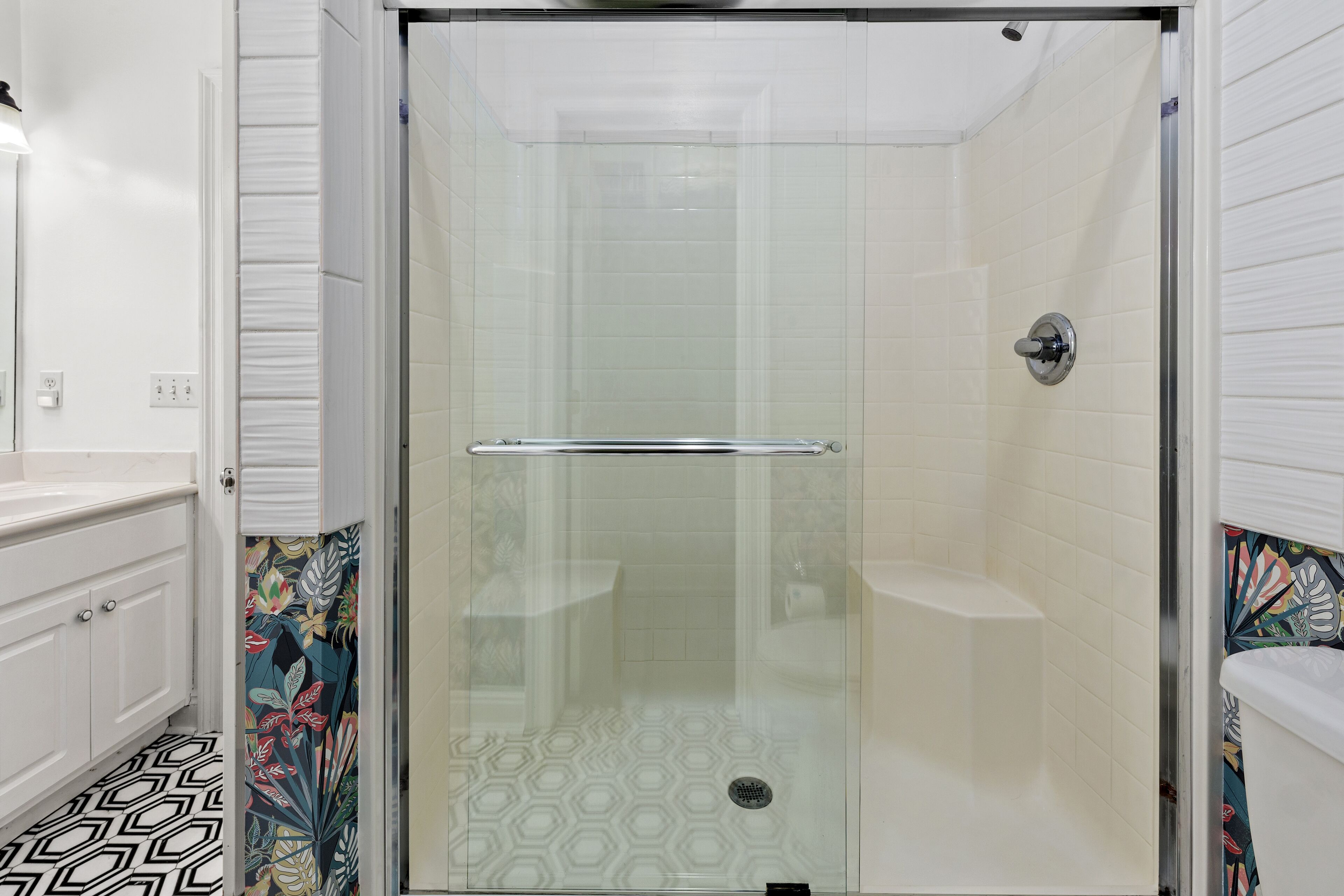 A bathroom with a glass-enclosed shower, white tiled walls, a white sink, and a patterned floor.