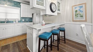 Une cuisine avec des meubles blancs, un pare-feu en carreaux bleus, un îlot central blanc avec des tabourets de bar et une horloge murale.