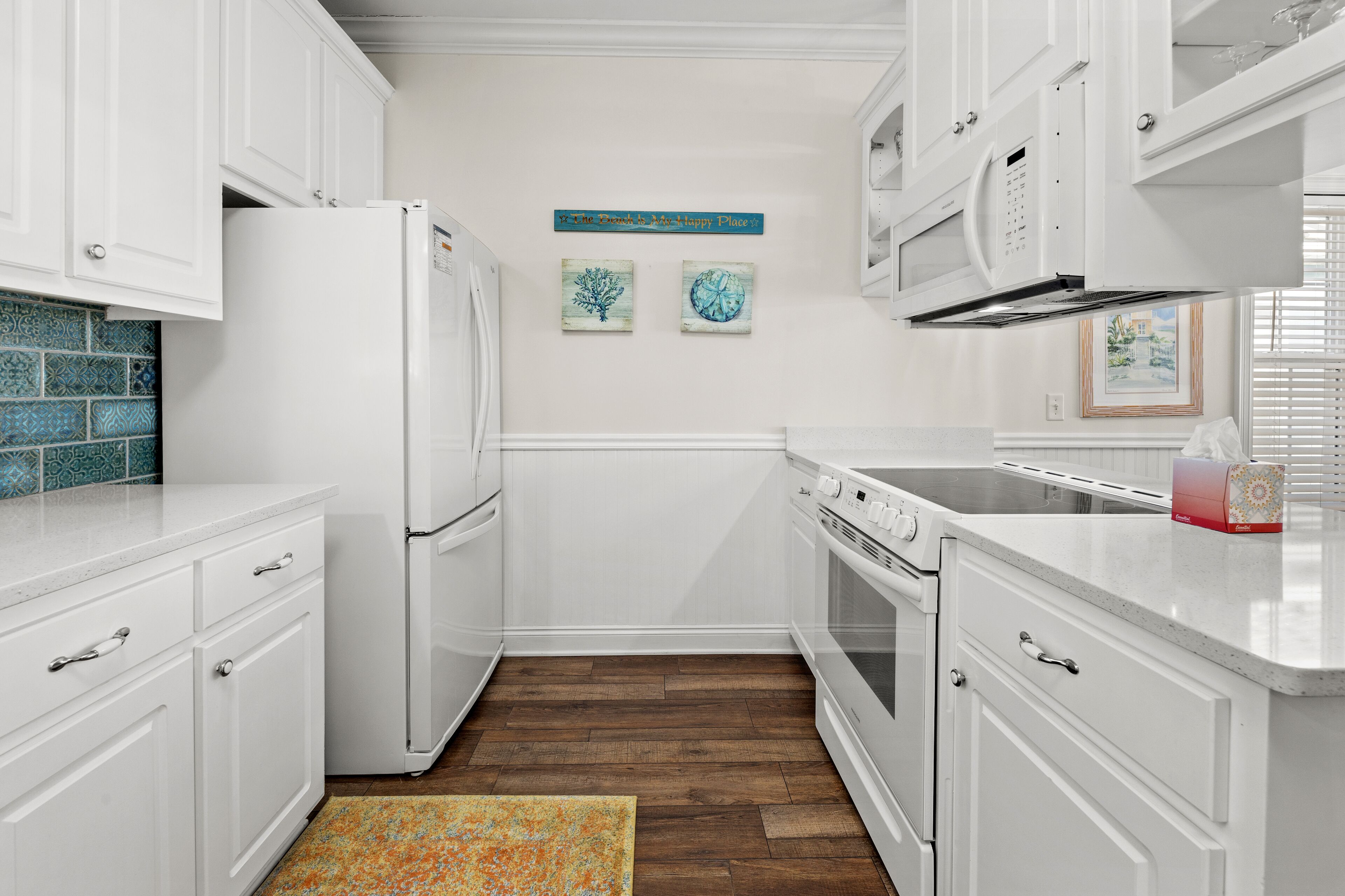 A kitchen with white cabinets, a refrigerator, a microwave, and a stove. There are two wall decorations and a rug on the floor.