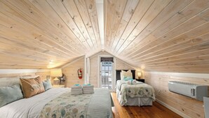 A cozy attic bedroom with wooden slats on the ceiling, two beds with matching bedding, a small table with a lamp, and a wall-mounted air conditioner. - Cute Beach Cottage Steps to the Beach! Panama City Beach, FL! (Panama City Beach)