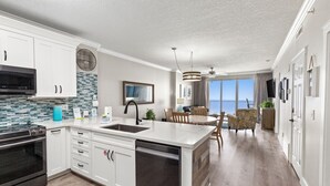 Eine moderne Küche mit weißen Schränken, einem Geschirrspüler und einer großen Kochinsel. Es gibt eine Essecke mit Tisch und Stühlen sowie einen Blick auf den Ozean durch große Fenster.