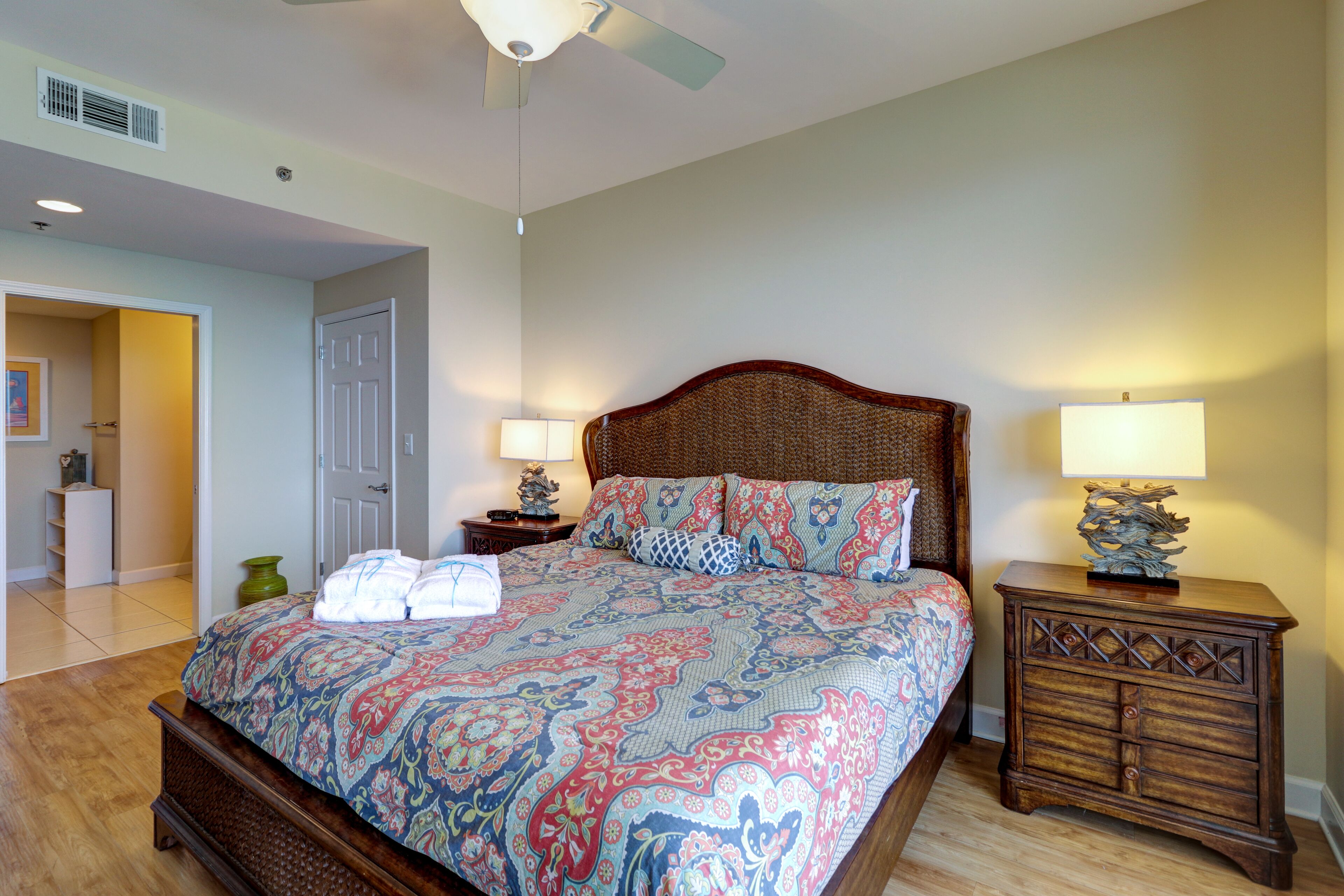 A bedroom with a large bed, two bedside tables with lamps, a ceiling fan, and a door leading to another room.
