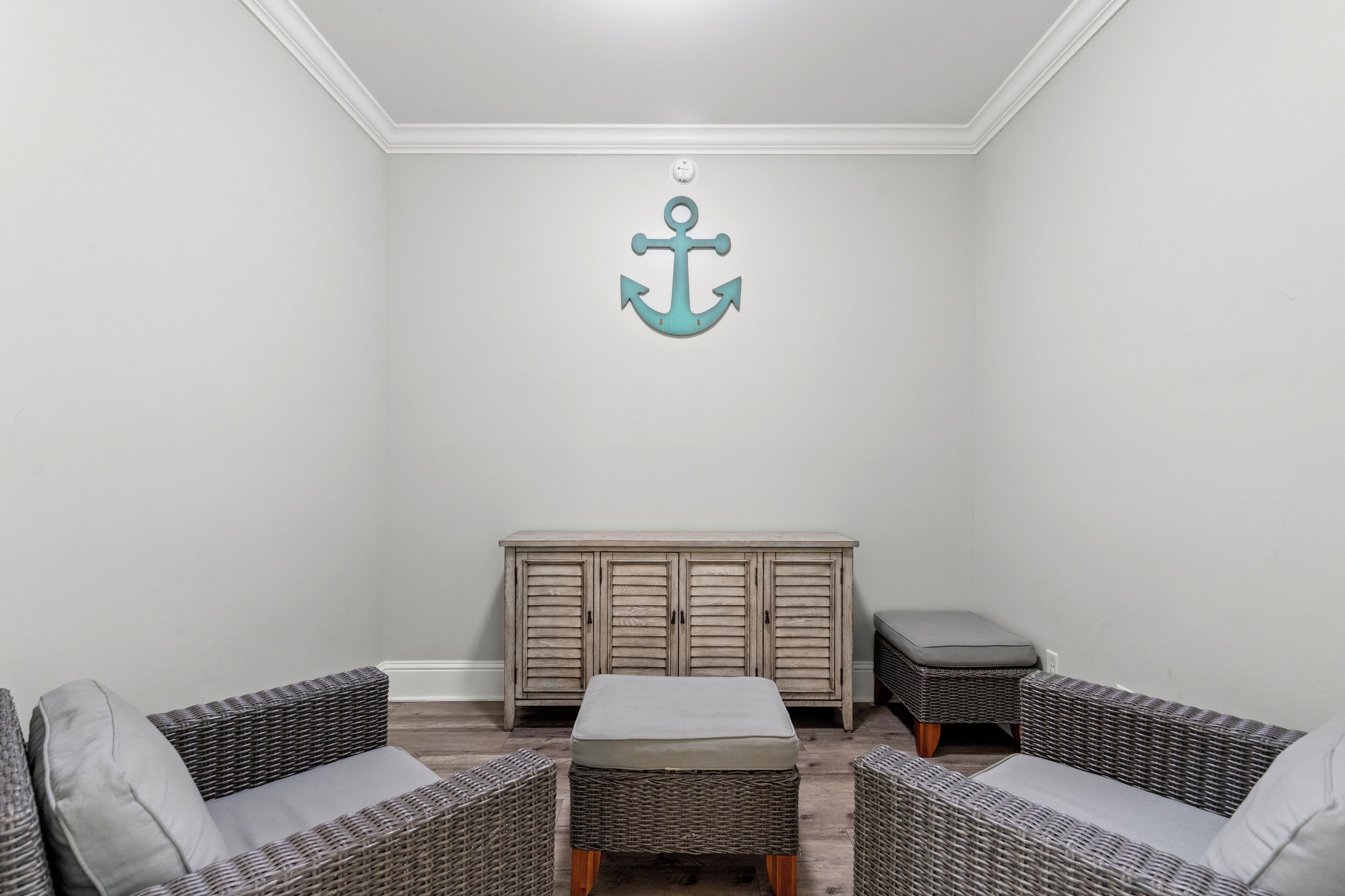 Una habitación con dos sofás de mimbre, un pequeño reposapiés y un mueble de madera frente a una pared blanca decorada con una ancla.