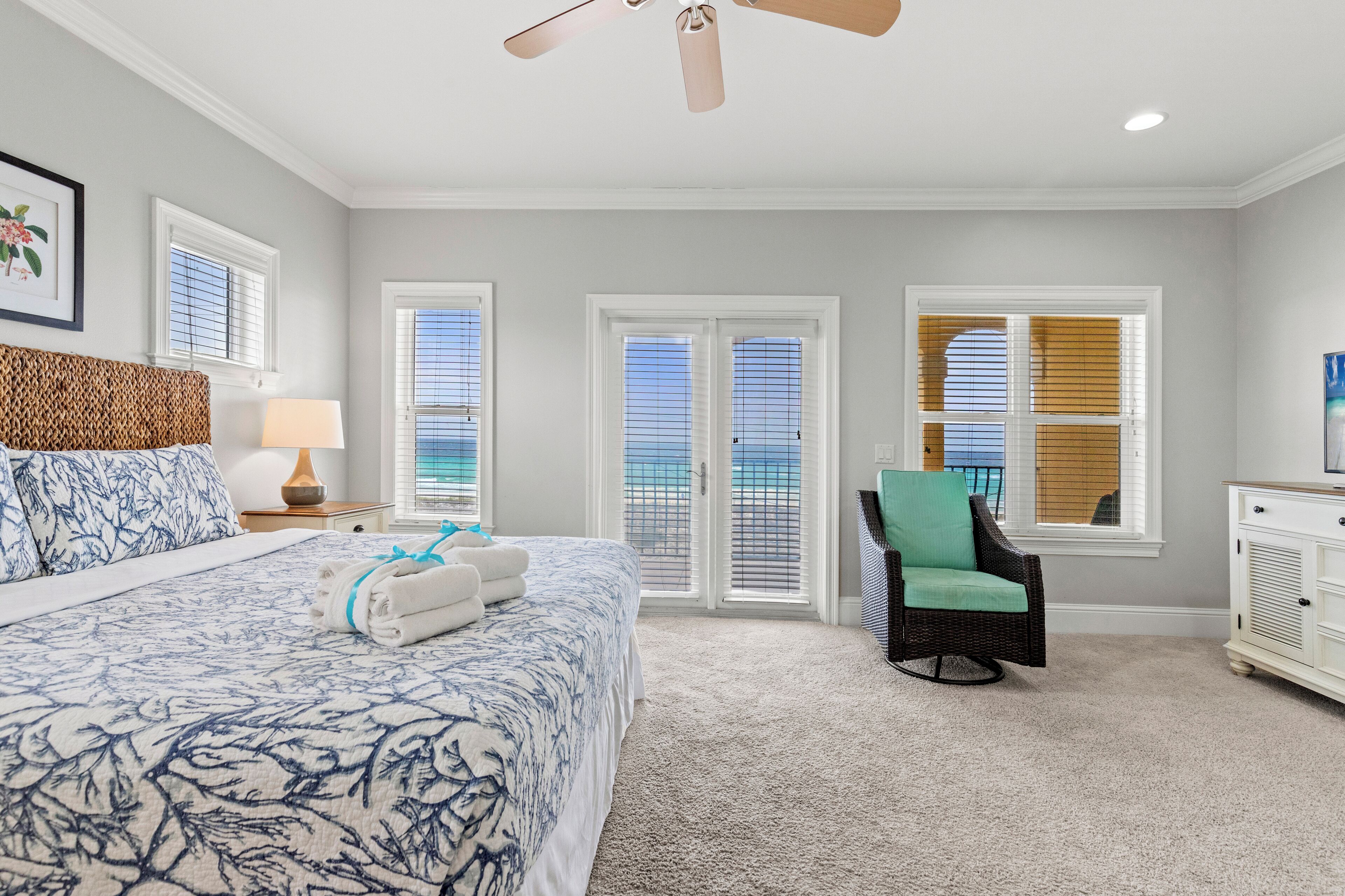 A bedroom with a large bed, two windows with blinds, a ceiling fan, and a view of the ocean.