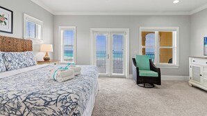 A bedroom with a large bed, two windows with blinds, a ceiling fan, and a view of the ocean.