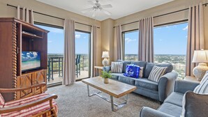 A spacious living room with a large TV, comfortable seating, a balcony with a view, and a ceiling fan.