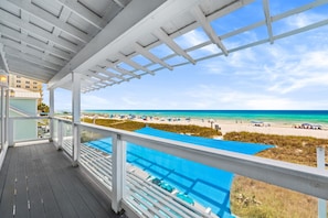 A covered balcony with a view of a beach and a pool.