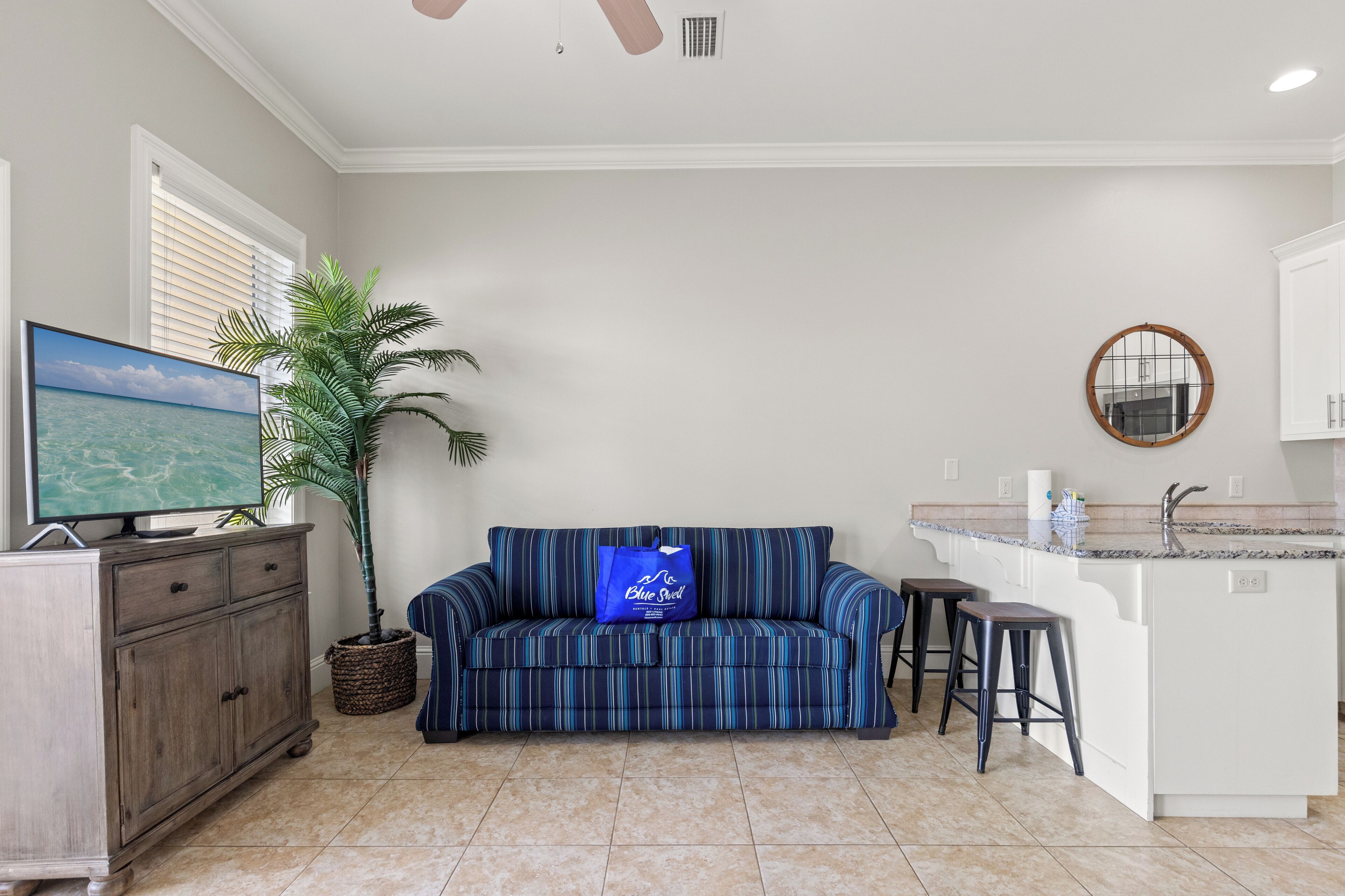 A living room with a TV, a sofa, a coffee table, a potted plant, and a kitchen area with a sink and stools.