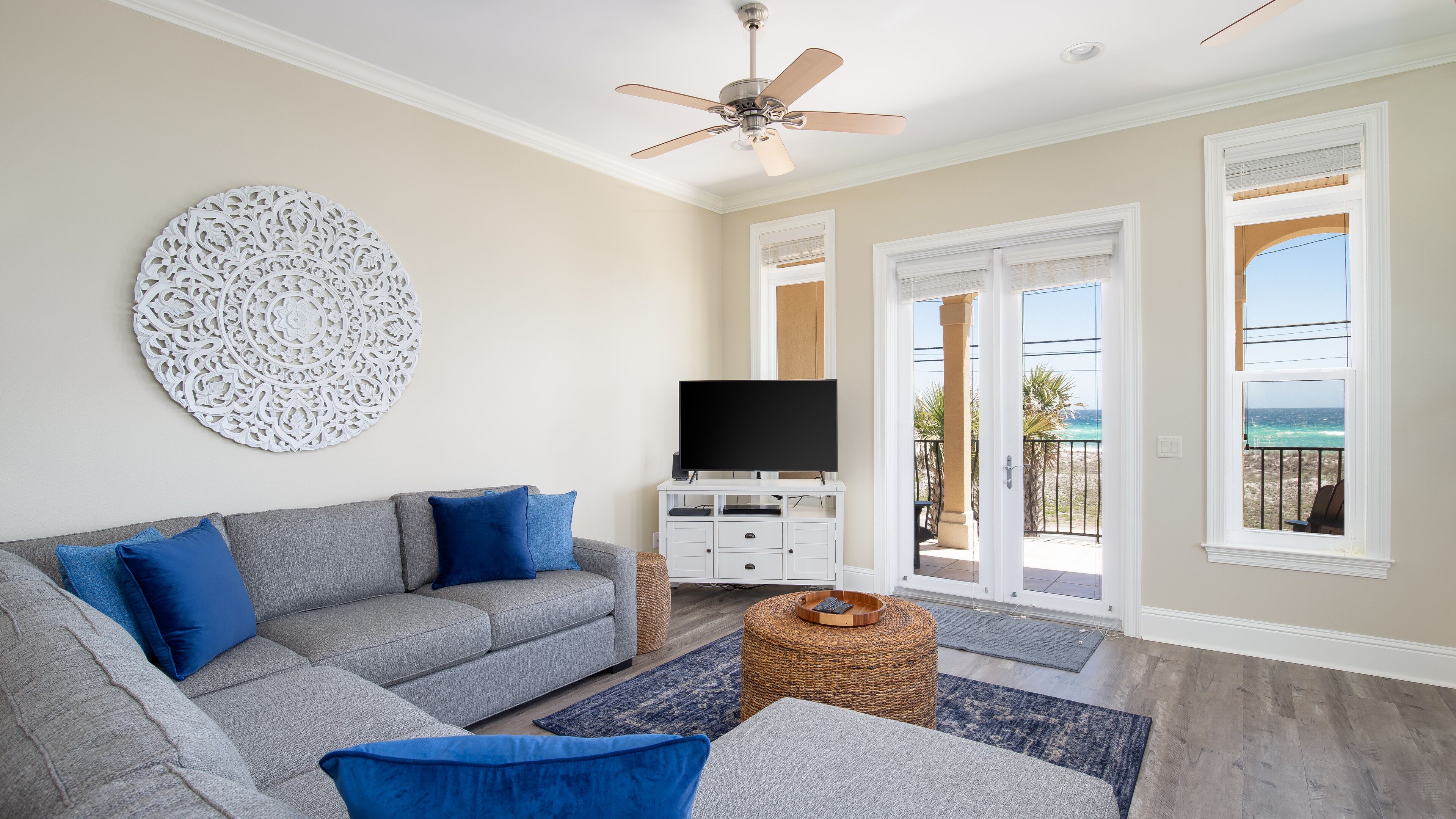 A living room with a grey sofa, blue cushions, a flat-screen TV, a decorative wall piece, and a view of the ocean through a balcony door.